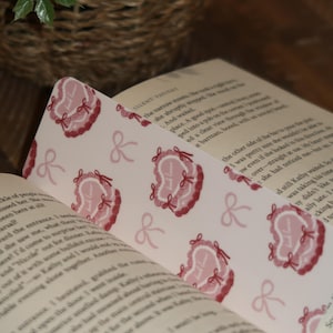May include: A bookmark with a repeating pattern of pink heart-shaped designs with the text "Just Love" and bows. The bookmark is placed in an open book with printed text.