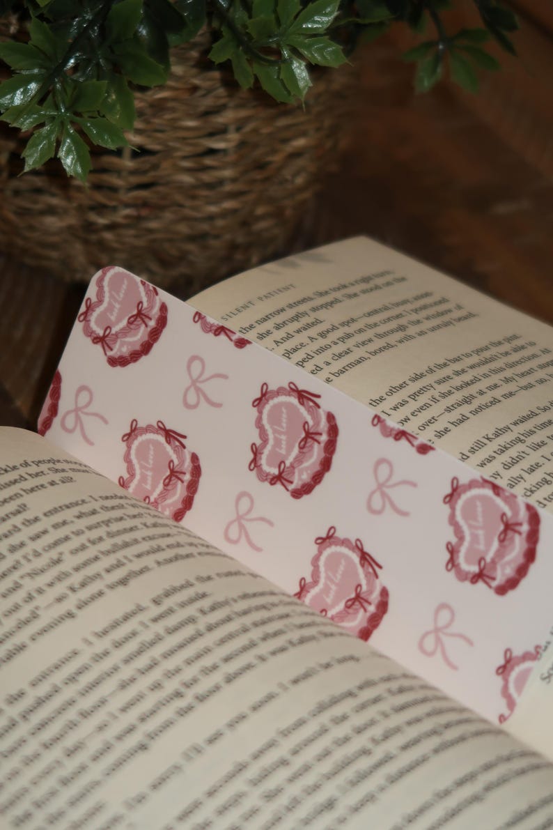 May include: A bookmark with a repeating pattern of pink heart-shaped designs with the text "Just Love" and bows. The bookmark is placed in an open book with printed text.