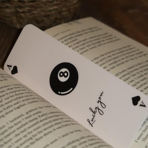May include: White bookmark with a black eight ball graphic and the text "Lucky you". The bookmark also features a black Ace of Spades symbol. The bookmark is placed inside an open book.