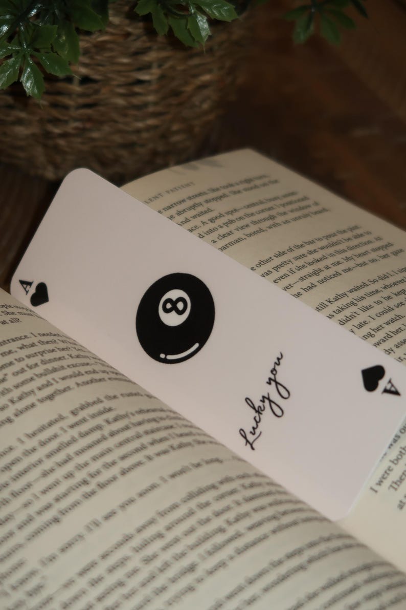 May include: White bookmark with a black eight ball graphic and the text "Lucky you". The bookmark also features a black Ace of Spades symbol. The bookmark is placed inside an open book.