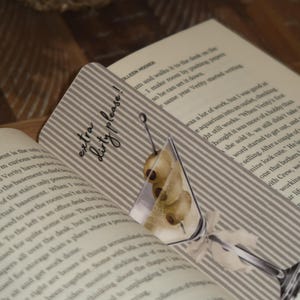 May include: A decorative bookmark featuring a martini glass illustration with olives. The bookmark has a striped background and the text "extra dirty please!". It is placed in an open book.