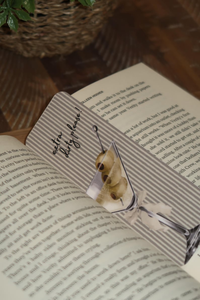 May include: A decorative bookmark featuring a martini glass illustration with olives. The bookmark has a striped background and the text "extra dirty please!". It is placed in an open book.