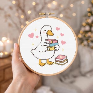May include: A cross-stitch embroidery hoop showing a cartoon goose carrying books, with pink hearts and sparkles. The goose has a yellow beak and feet. The hoop is held in a hand, with a blurred background of lights.