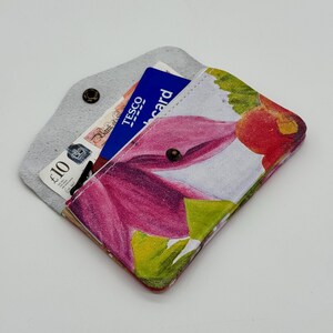 May include: A small, open wallet with a floral design. The wallet is open, revealing a blue Tesco card and a ten-pound note. The floral pattern features pink, red, and green hues. The wallet is made of a soft material and has a snap closure.