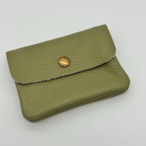 May include: A small, olive green leather wallet with a textured surface. The wallet has a flap closure secured by a gold-colored snap. The edges of the flap are raw and unfinished.