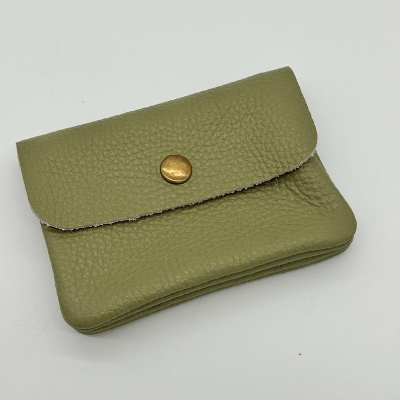 May include: A small, olive green leather wallet with a textured surface. The wallet has a flap closure secured by a gold-colored snap. The edges of the flap are raw and unfinished.