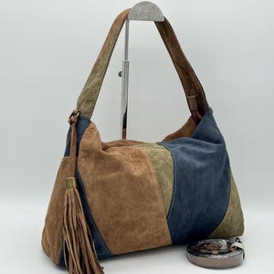 May include: A patchwork suede shoulder bag in shades of brown, green, and blue. The bag has a long strap, a tassel, and a small belt. The bag is displayed on a white background.