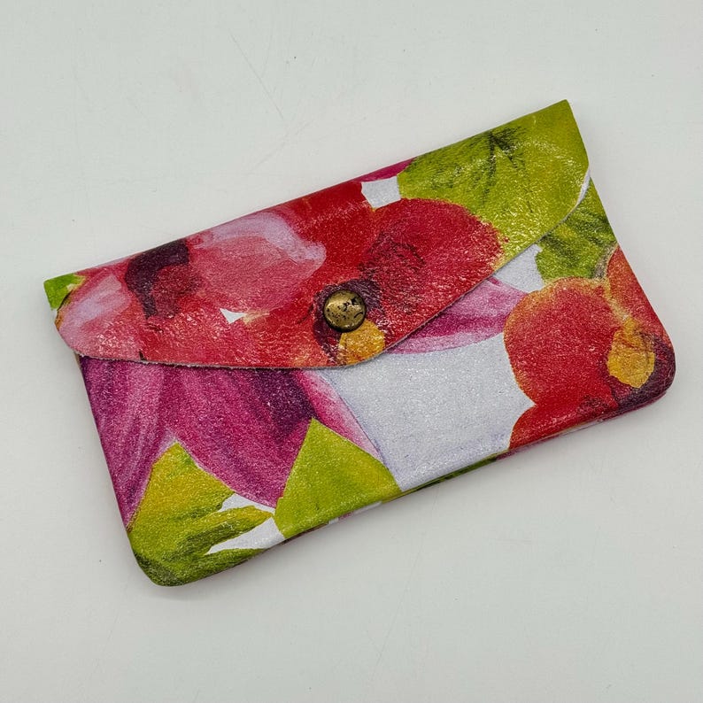 May include: A floral print card holder with a brass snap closure. The wallet features a vibrant pattern of red, pink, and green flowers on a white background. The card holder is made of a durable material.