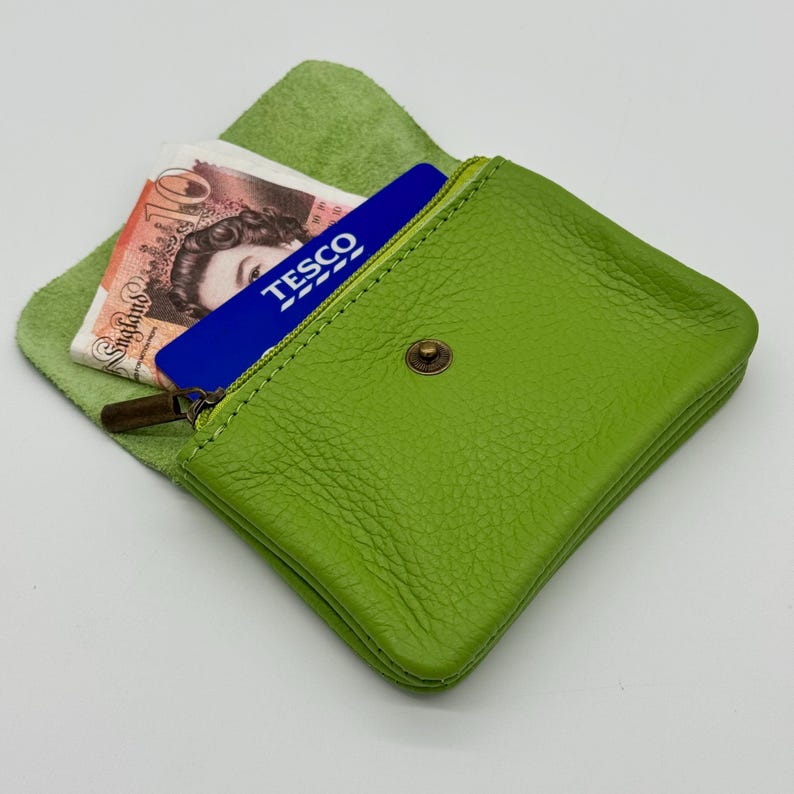 May include: A lime green leather coin purse with a zipper and a snap closure. The purse is open, revealing a blue Tesco card and a British ten-pound note. The purse is on a light green leather background.