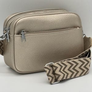 May include: A gold-coloured crossbody bag with a textured finish. The bag has two zipped compartments and a patterned strap with silver-tone hardware. The strap features a brown and beige zigzag design.