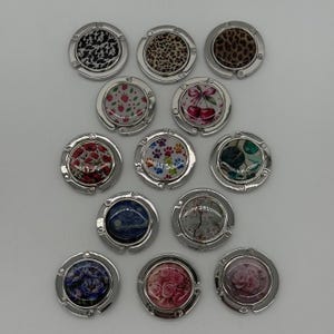 May include: A collection of twelve circular metal bag hooks, each featuring a unique decorative insert. Designs include animal prints, strawberries, cherries, floral patterns, paw prints, and artistic motifs, all set within polished silver-toned frames.