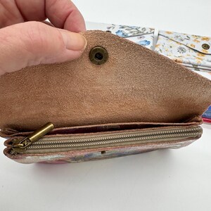 May include: Open view of a small, floral-patterned wallet with a brown leather flap secured by a brass snap. The interior reveals a zippered compartment and a floral print. Additional floral wallets are visible in the background.