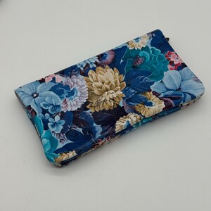 May include: A rectangular clutch with a floral pattern in shades of blue, teal, and beige. The flowers are large and detailed, set against a dark blue background. The clutch has a zipper closure and a soft, textured appearance.