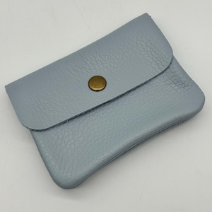 May include: A small, light blue leather wallet with a flap closure and a brass-colored button. The wallet has a textured surface and a rounded shape. The design is simple and minimalist.