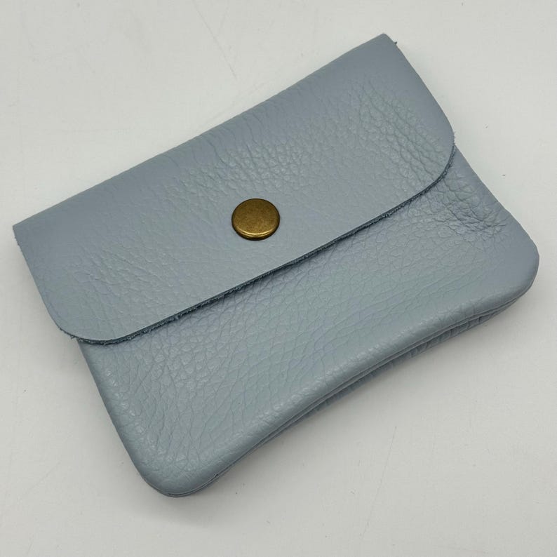 May include: A small, light blue leather wallet with a flap closure and a brass-colored button. The wallet has a textured surface and a rounded shape. The design is simple and minimalist.