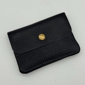 May include: A black leather wallet with a textured surface. The wallet has a flap closure secured by a gold-colored button. The wallet is rectangular and appears to be empty.