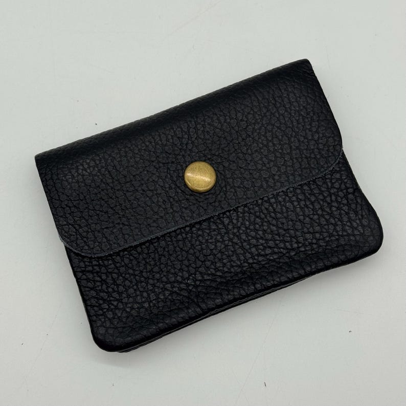 May include: A black leather wallet with a textured surface. The wallet has a flap closure secured by a gold-colored button. The wallet is rectangular and appears to be empty.