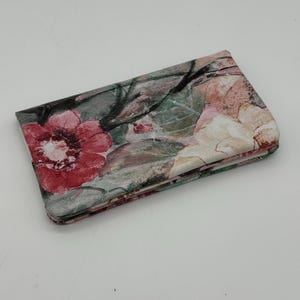 May include: Folded floral wallet featuring a watercolor design with red, pink, and cream-colored flowers. The wallet has a green and gray background with black accents. The wallet is folded and appears to be made of fabric.