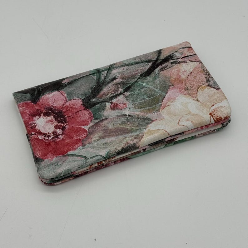 May include: Folded floral wallet featuring a watercolor design with red, pink, and cream-colored flowers. The wallet has a green and gray background with black accents. The wallet is folded and appears to be made of fabric.