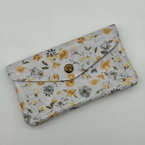 May include: A light gray, floral-patterned wallet with a flap closure and a brass-colored button. The wallet features a repeating pattern of yellow and gray flowers and leaves. The wallet is closed.
