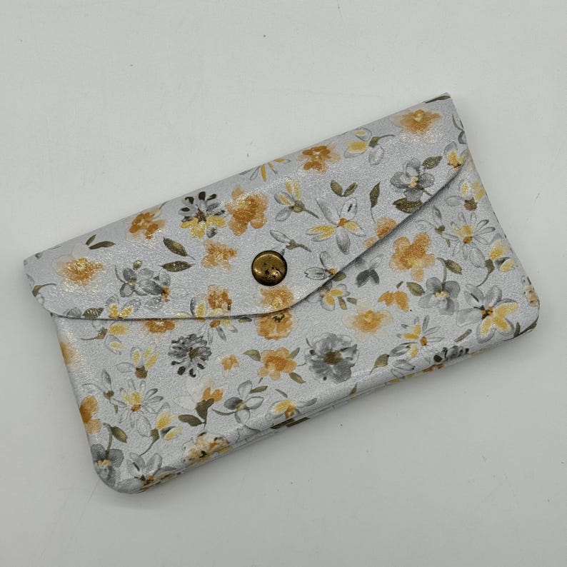 May include: A light gray, floral-patterned wallet with a flap closure and a brass-colored button. The wallet features a repeating pattern of yellow and gray flowers and leaves. The wallet is closed.