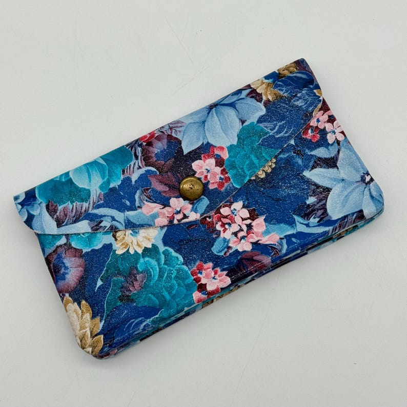 May include: A blue floral clutch with a brass button closure. The clutch features a pattern of blue, pink, and cream-colored flowers on a dark blue background. The clutch is rectangular and has a flap closure.