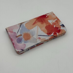 May include: A floral print wallet featuring watercolor-style flowers in shades of red, orange, pink, and blue. The wallet is rectangular and folded, with a soft, textured appearance.