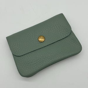 May include: A sage green leather cardholder with a textured surface. The cardholder has a flap closure secured by a round, brass-colored button. The item is rectangular and appears to be empty.
