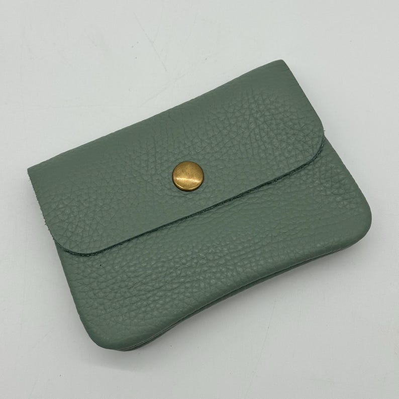May include: A sage green leather cardholder with a textured surface. The cardholder has a flap closure secured by a round, brass-colored button. The item is rectangular and appears to be empty.