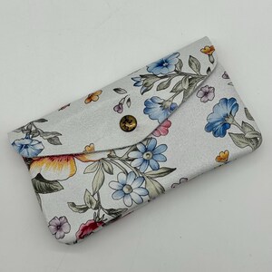 May include: A white floral print card holder with a snap closure. The design features blue, pink, and yellow flowers with green leaves. The card holder is rectangular and has a button closure.