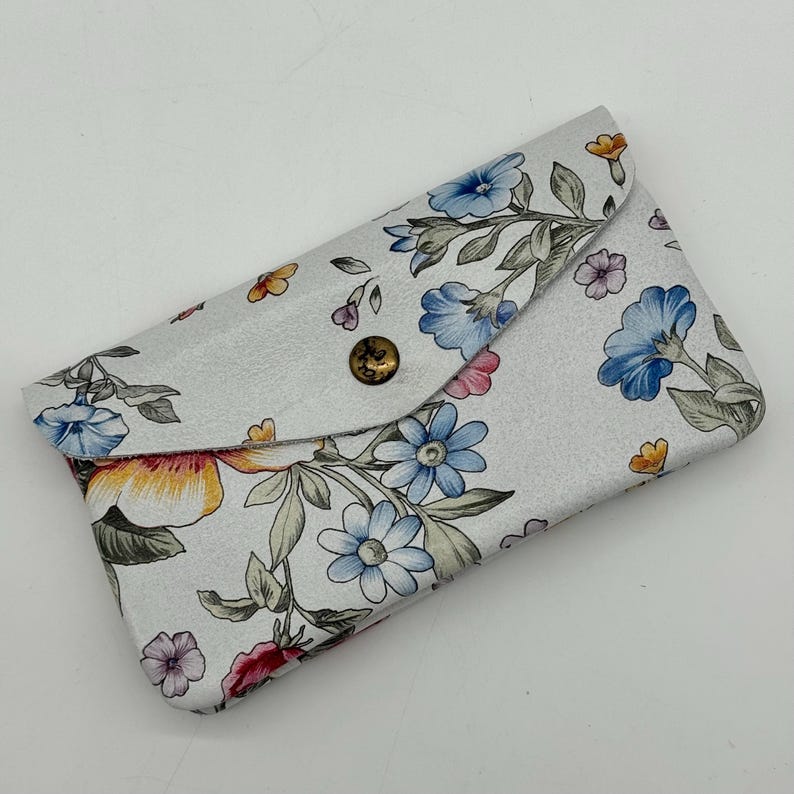 May include: A white floral print card holder with a snap closure. The design features blue, pink, and yellow flowers with green leaves. The card holder is rectangular and has a button closure.