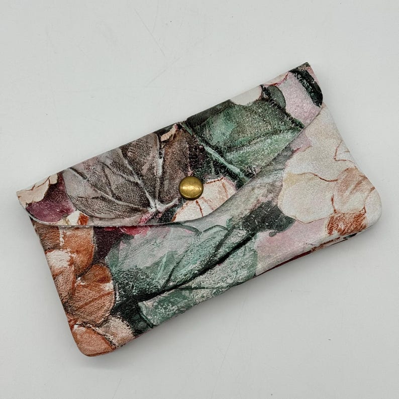 May include: A floral print clutch with a brass snap closure. The clutch features a pattern of muted colors, including brown, green, and pink, resembling a watercolor painting of flowers and leaves. The clutch is closed with a round, gold-colored snap.
