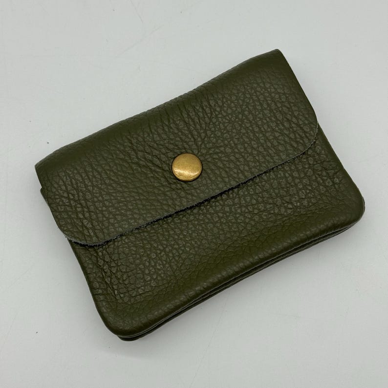 May include: Olive green leather wallet with a textured surface. The wallet has a flap closure secured by a gold-colored button. The wallet is rectangular and appears to be empty.