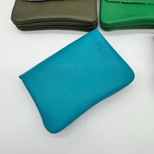 May include: A close-up shot of several small, rectangular leather pouches in various colors. The primary focus is on a vibrant teal pouch with a textured surface. Other pouches are olive green and bright green.