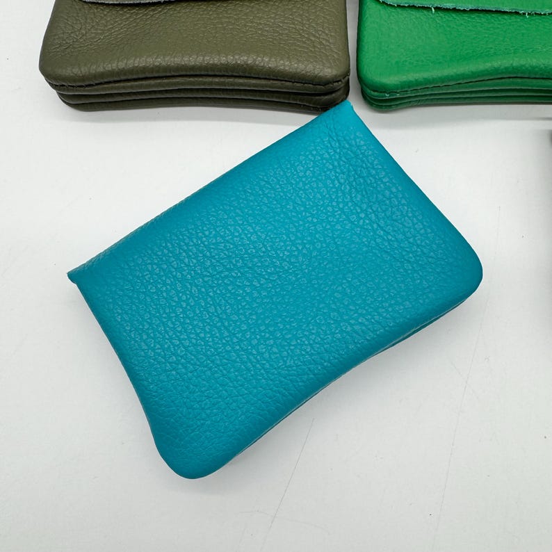 May include: A close-up shot of several small, rectangular leather pouches in various colors. The primary focus is on a vibrant teal pouch with a textured surface. Other pouches are olive green and bright green.