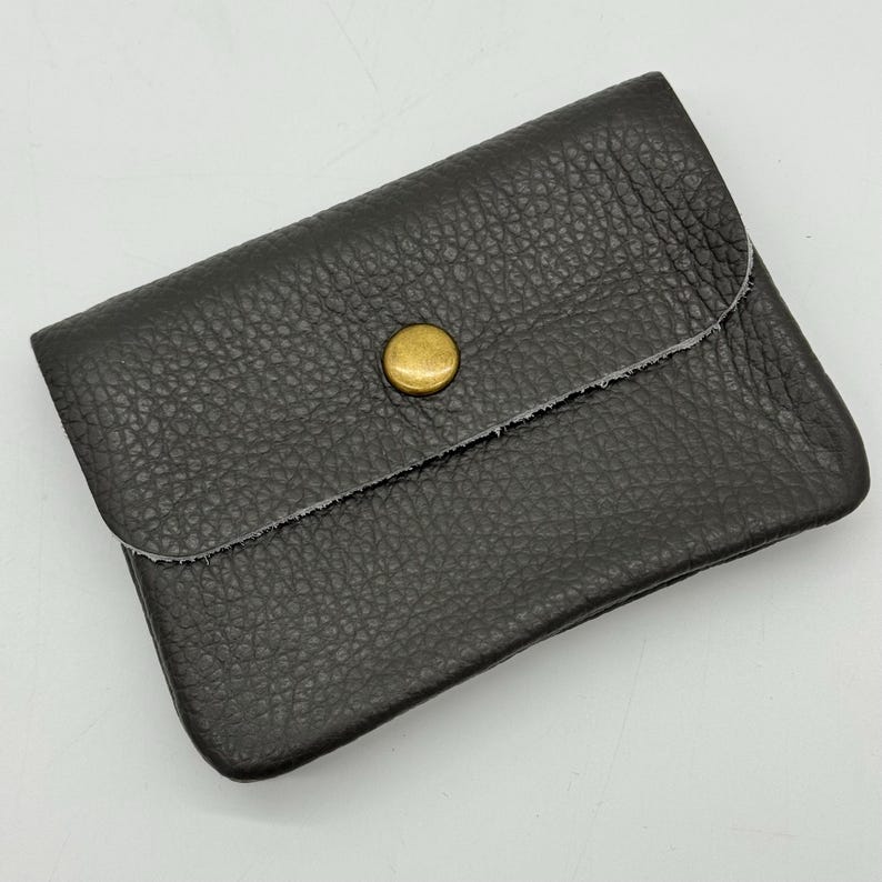 May include: A dark gray leather wallet with a textured surface. The wallet has a flap closure secured by a gold-colored button. The edges of the wallet are raw and unfinished.