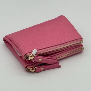 K&ouml;nnte beinhalten: Ein leuchtend pinkfarbenes Portemonnaie mit strukturierter Oberfl&auml;che und zwei horizontalen Rei&szlig;verschlussf&auml;chern. Jeder Rei&szlig;verschluss hat einen passenden pinkfarbenen Zipper mit einem kleinen goldfarbenen Metallniet. Das Portemonnaie ist gefaltet.