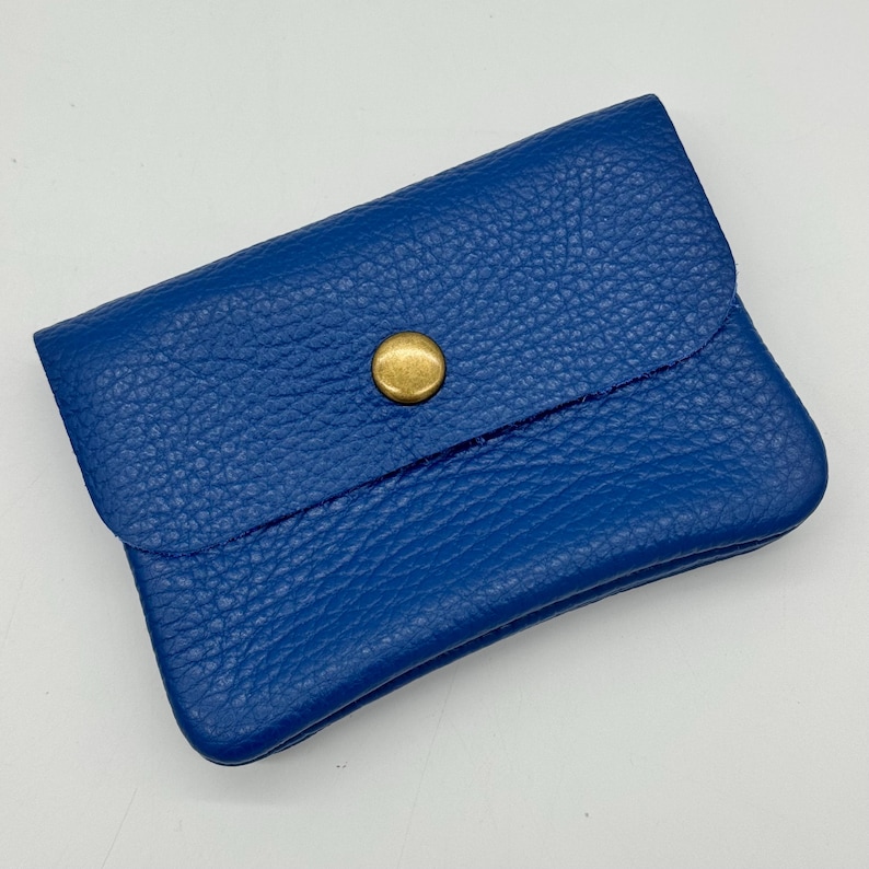 May include: A small, cobalt blue leather wallet with a textured surface. The wallet has a flap closure secured by a gold-colored snap. The wallet is rectangular and appears to be empty.