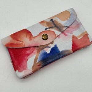 May include: A small, rectangular leather cardholder with a watercolor floral design. The design features red, orange, blue, and brown hues on a white background. The cardholder has a flap closure secured by a brass button.