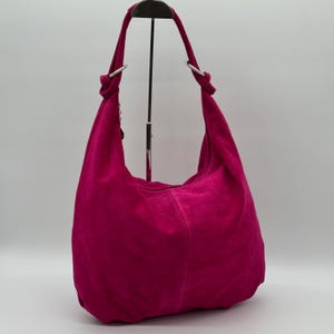 May include: A fuchsia suede hobo bag with a single shoulder strap. The bag has a rounded shape and is suspended from a metal stand. The strap is attached to the bag with silver-coloured metal rings.