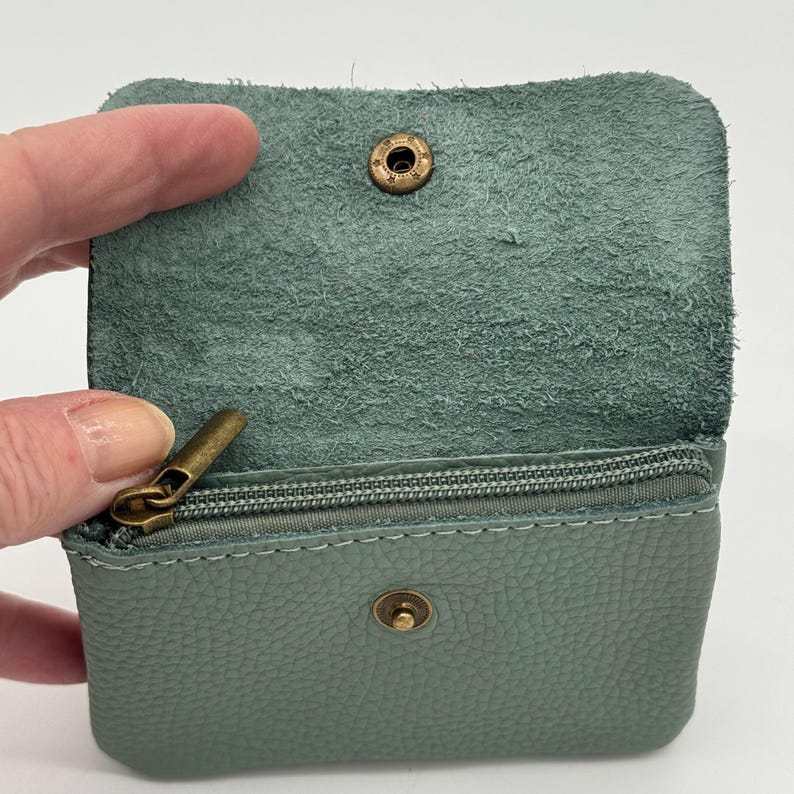 May include: Open, sage green leather coin purse with a zipper closure and a snap closure. The interior is a lighter shade of green. The purse has a textured exterior and a brass-colored zipper and snap.