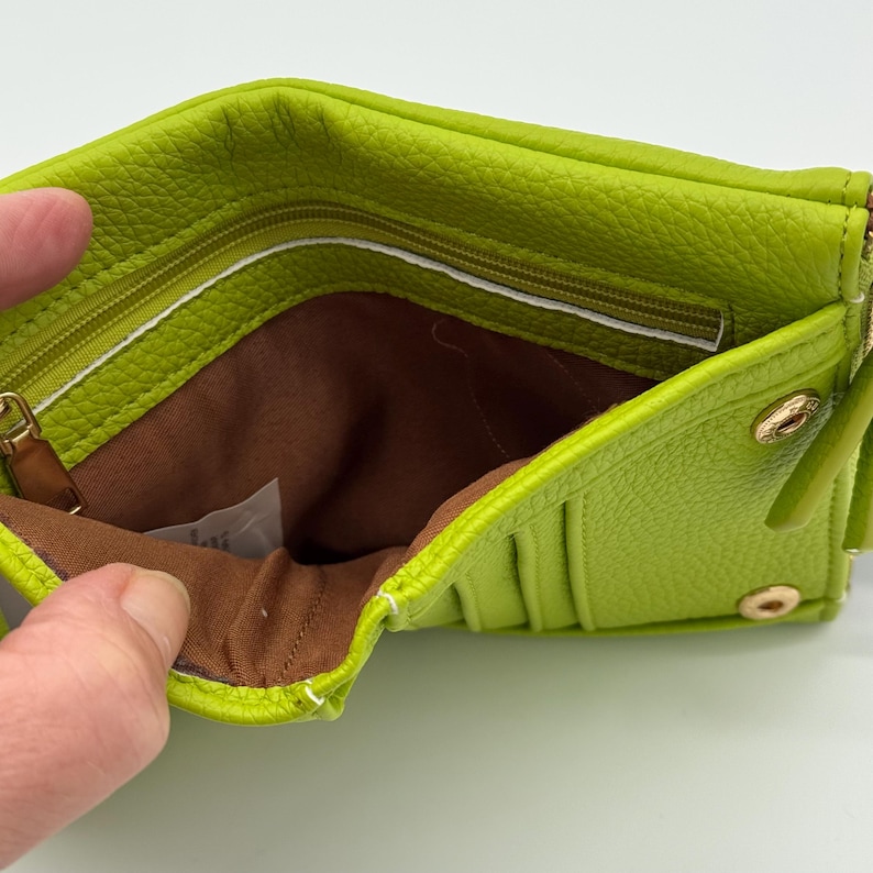 K&ouml;nnte beinhalten: Eine limettengr&uuml;ne Geldb&ouml;rse aus strukturiertem Leder mit braunem Stoffinnenfutter, einem Rei&szlig;verschlussfach und mehreren Kartenf&auml;chern. Die Geldb&ouml;rse wird mit goldfarbenen Metalldruckkn&ouml;pfen verschlossen und hat einen passenden Rei&szlig;verschluss.