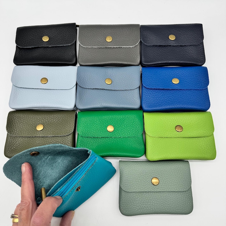 May include: A collection of small leather coin purses in various colors, including black, gray, blue, green, and teal. Each purse has a snap closure and a gold-colored button. One purse is open, revealing a teal interior.