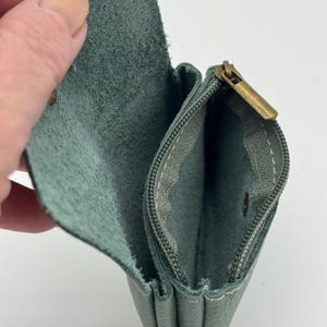 May include: Open view of a teal leather wallet with a brass zipper and snap closure. The interior is a lighter shade of teal, with visible stitching. The wallet is held open, showcasing its compartments.