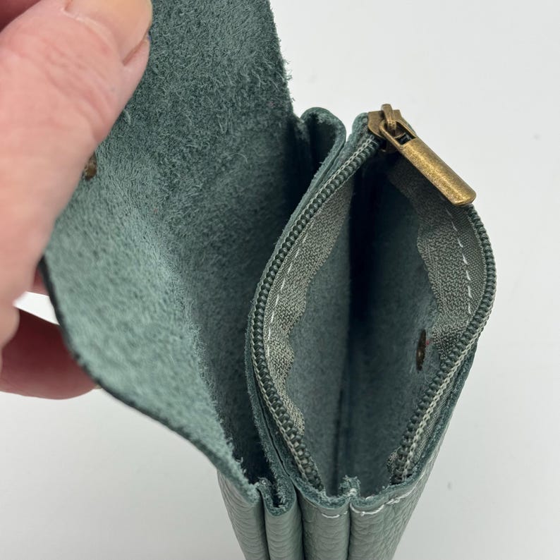 May include: Open view of a teal leather wallet with a brass zipper and snap closure. The interior is a lighter shade of teal, with visible stitching. The wallet is held open, showcasing its compartments.