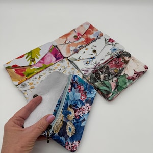 May include: A collection of floral print wallets in various colors. One wallet is open, revealing a light-colored interior. The wallets feature a snap closure and are arranged on a white surface. The floral patterns include pink, blue, and yellow.