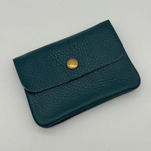 May include: Teal leather wallet with a textured surface and a flap secured by a gold-colored button. The wallet is rectangular and appears to be closed. The leather has a pebbled texture.