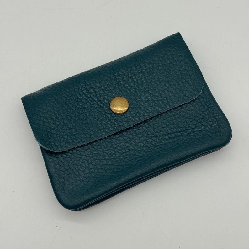 May include: Teal leather wallet with a textured surface and a flap secured by a gold-colored button. The wallet is rectangular and appears to be closed. The leather has a pebbled texture.