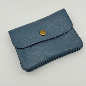May include: A small, blue-grey leather wallet with a textured surface. The wallet has a flap closure secured by a round, brass-colored button. The edges are neatly stitched with dark thread. The wallet is designed to be compact and portable.
