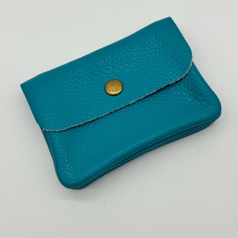 May include: A turquoise leather wallet with a textured surface. The wallet has a flap closure secured by a gold-colored button. The wallet is rectangular and appears to be empty.